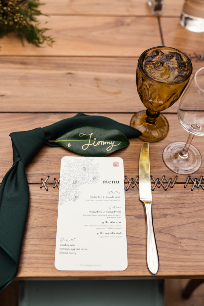 Groom's place setting wedding menu, green custom napkin, wine glass, and amber goblet on a wooden table for wedding reception at The Joinery in Chicago