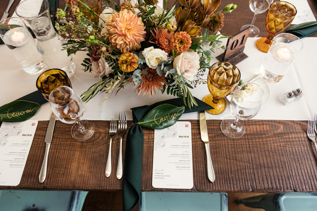 Guest table setting with floral centerpiece, menus, amber glasses, and green napkins labeled with names for wedding reception at The Joinery in Chicago