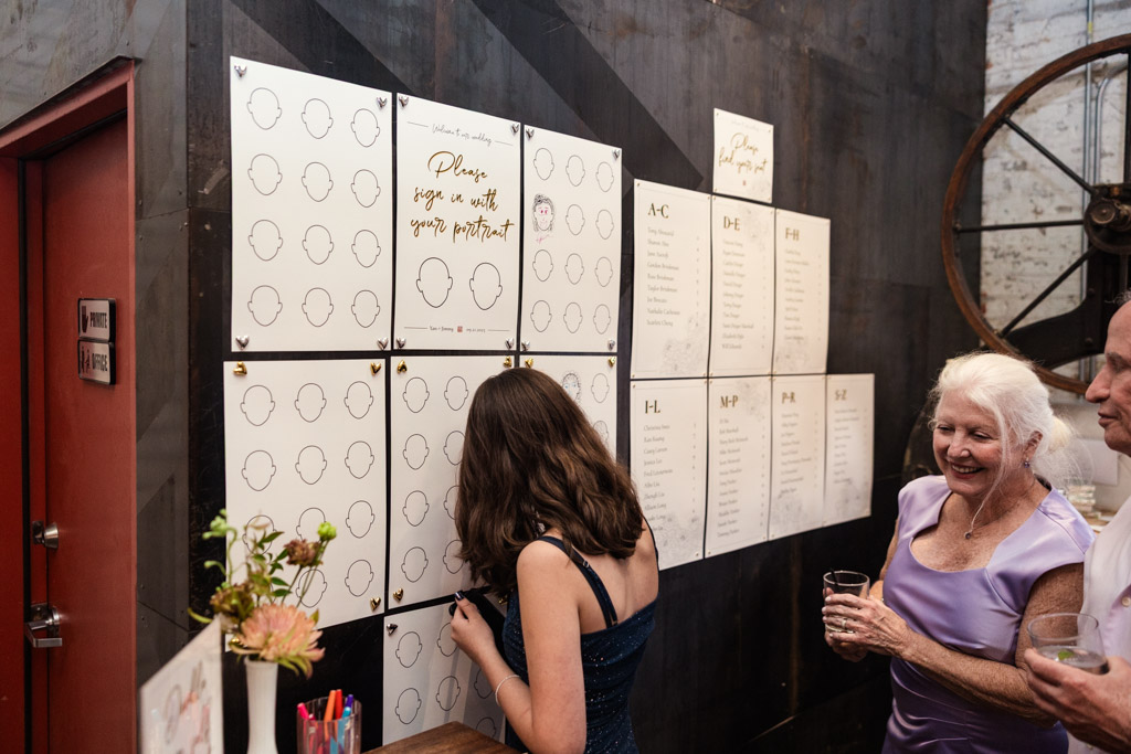 A guest signs a portrait wall at a Joinery wedding reception while two guests, smiling, watch nearby