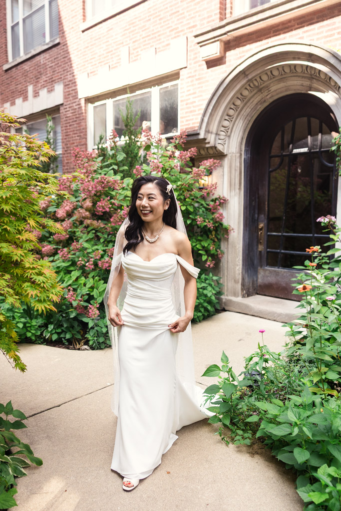 A smiling bride in a white dress walks outside a brick building during her first look
