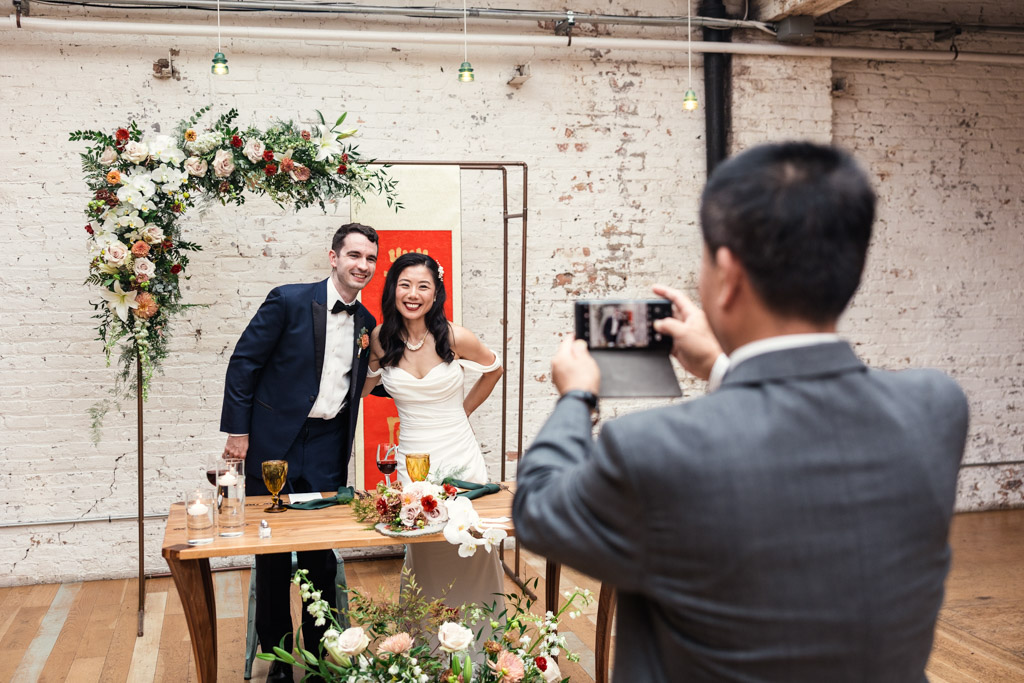 A couple poses and smiles at their wedding table while bride's father snaps their photo with a phone