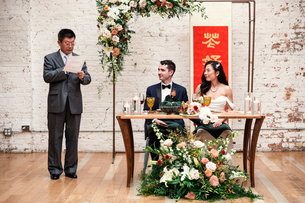 Father of the bride gives a speechwith bride and groom seated at a table adorned with flowers, with a red banner behind them