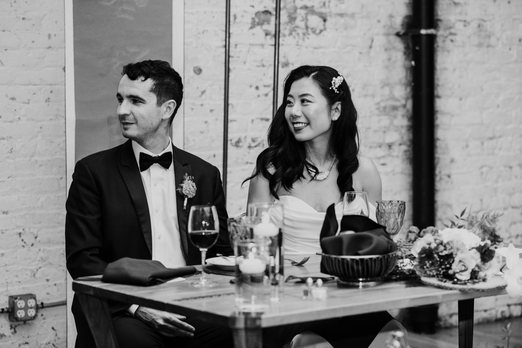 Black and white photo of bride and groom listening to speeches at a decorated table during their wedding reception at The Joinery in Chicago