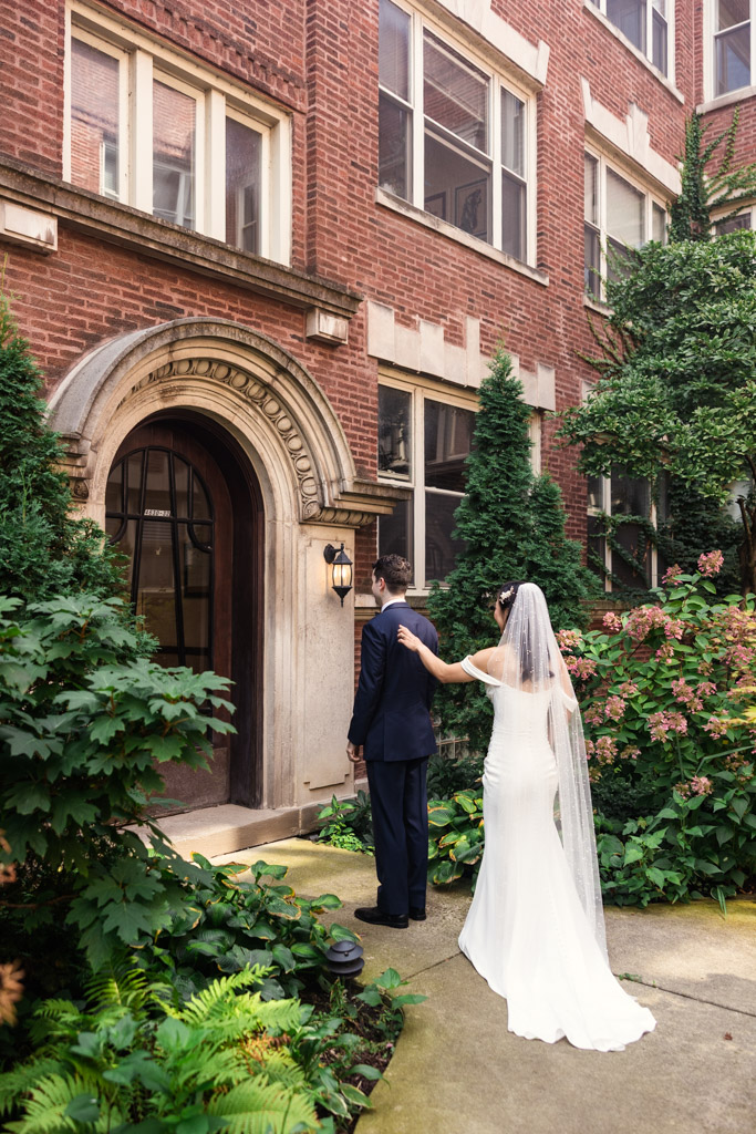 A bride in white touches the groom’s shoulder during their first look outside a brick building