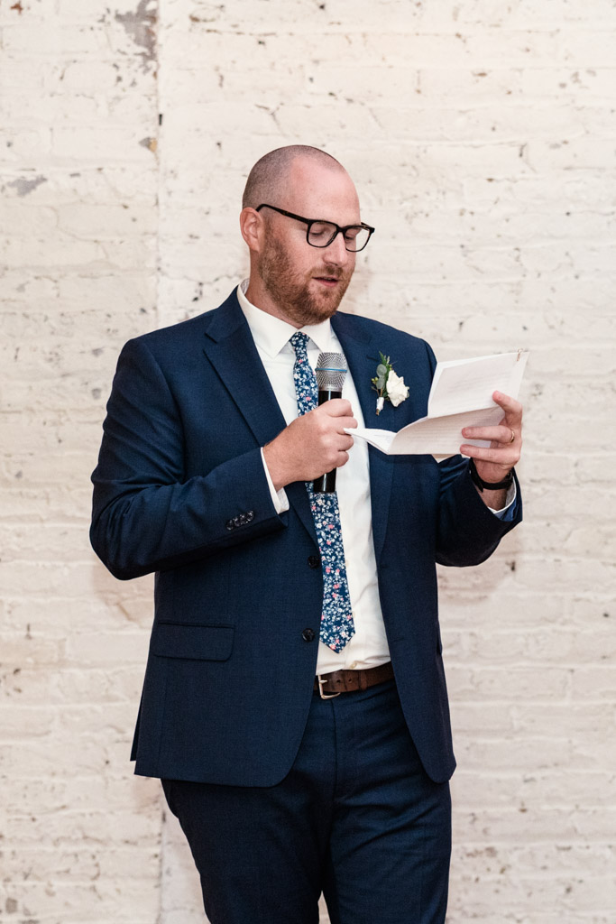 Man in a navy suit and glasses gives speech during wedding reception at The Joinery in Chicago