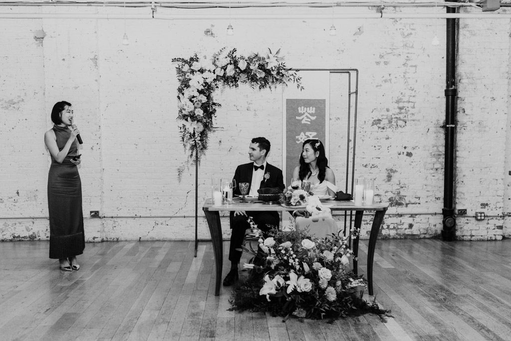 Black and white photo of woman giving a speech while the bride and groom listen from a decorated table during wedding reception at The Joinery in Chicago