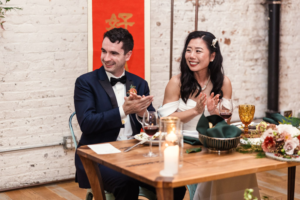Bride and groom smile and clap while seated at their head table during their wedding reception at The Joinery