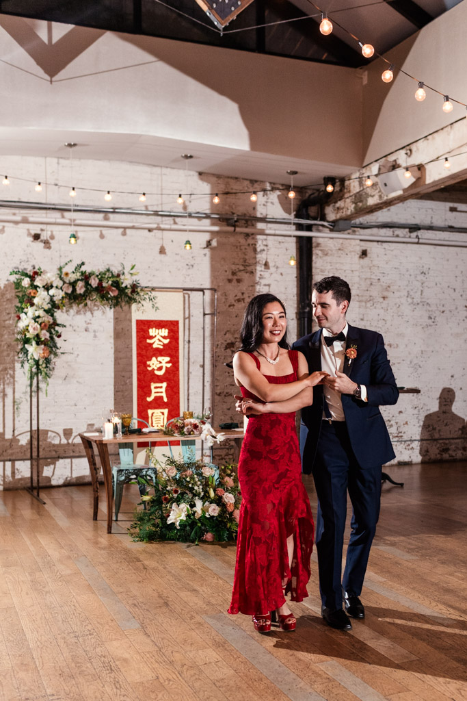 Happy newlywed couple dances during their wedding reception at The Joinery, floral arrangements, and a red Chinese banner in the background