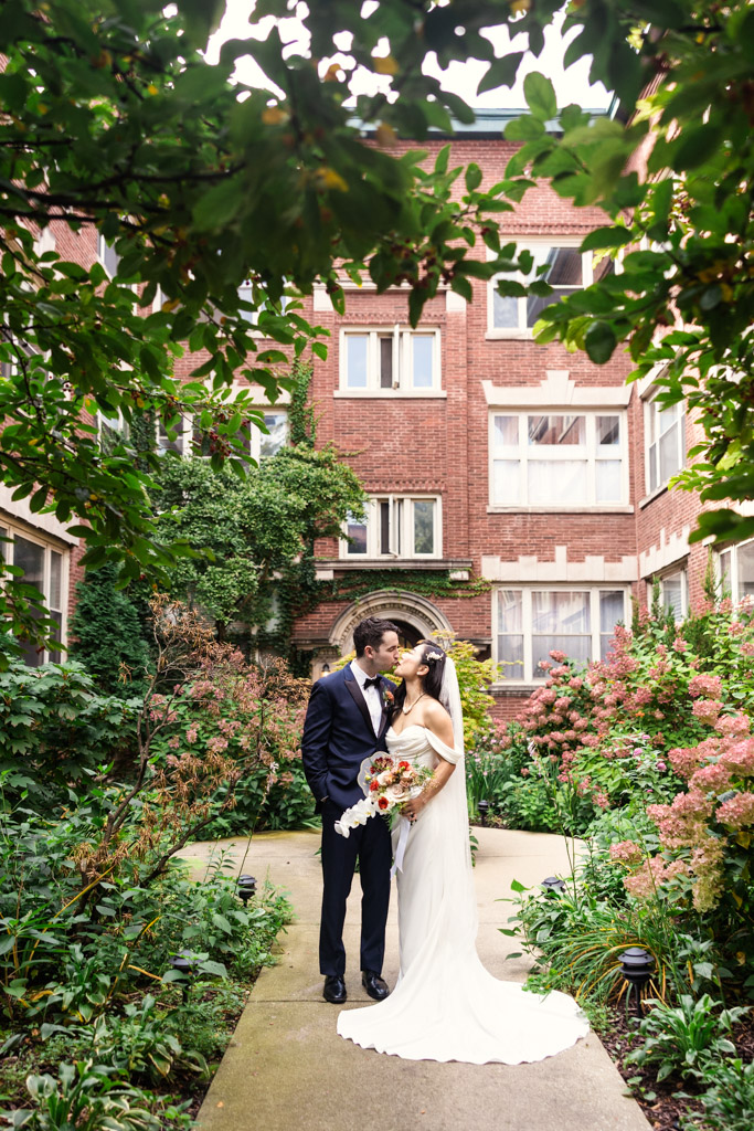 Bride and groom share a kiss in a garden courtyard, surrounded by lush greenery and the elegant joinery of a classic brick building.