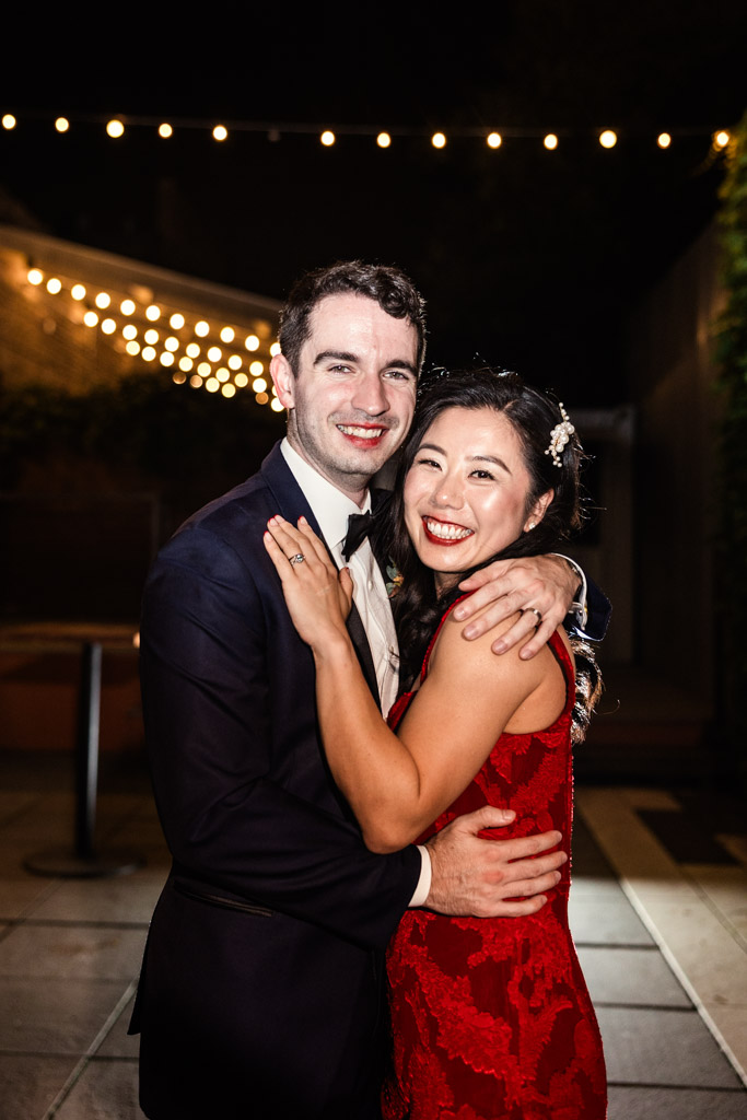 Portrait of newlywed couple in formal attire hugs and smiles at night under string lights outside The Joinery in Chicago