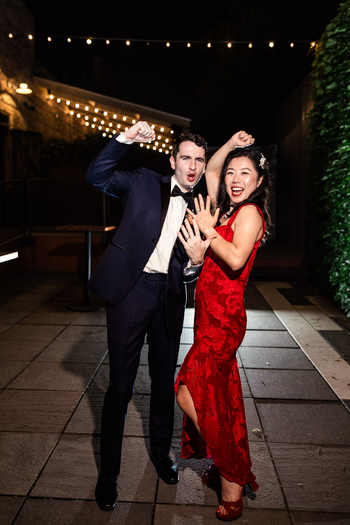 Groom in a tuxedo and bride in a red dress pose joyfully under string lights at night outside The Joinery wedding venue in Chicago