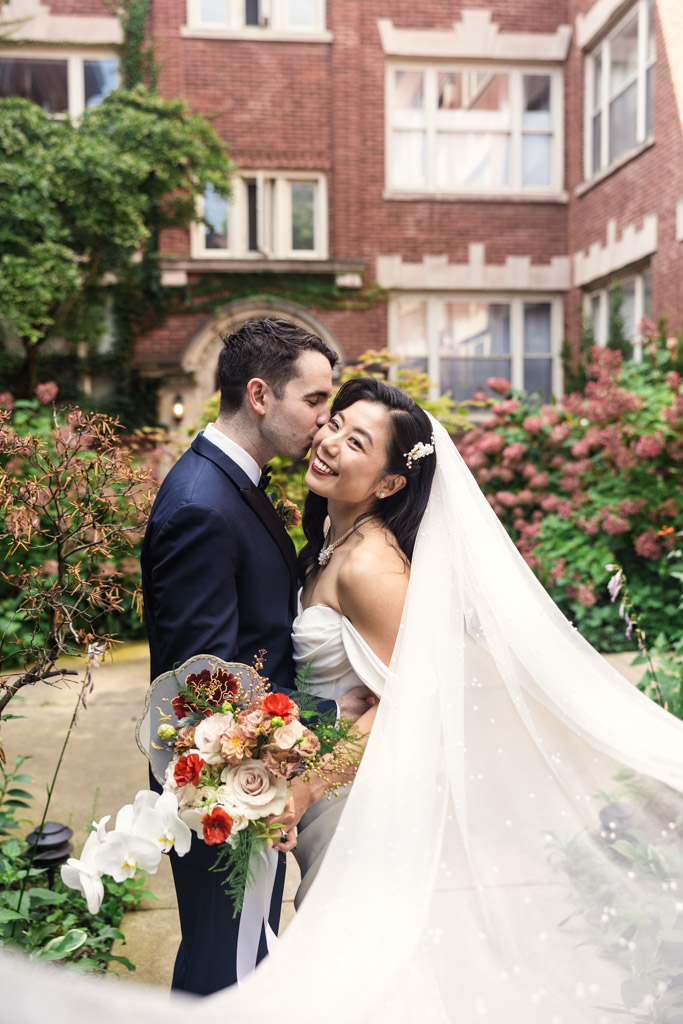 A bride and groom smile in a garden courtyard, the bride's veil flows behind her