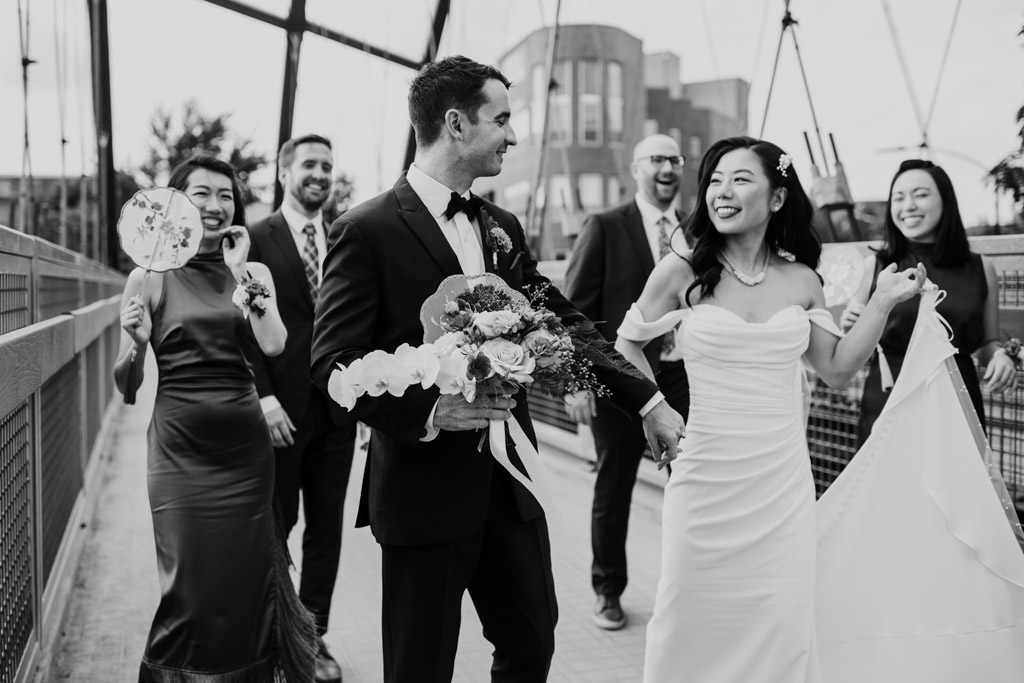 Black and white photo of bride and groom walking on the 606 Bridge, smiling and holding hands, with their wedding party behind them
