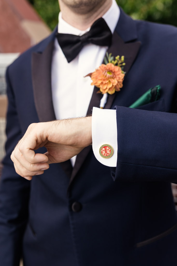 Groom in a navy tuxedo displays a cufflink with a red double happiness symbol