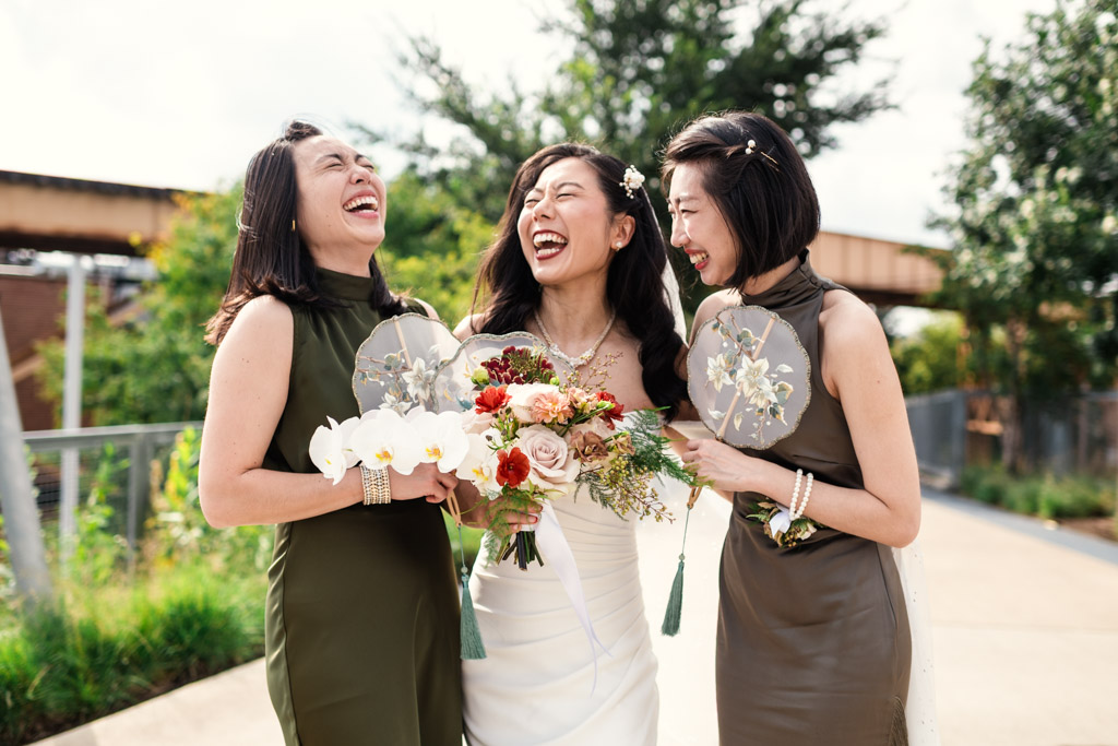 Bride and bridesmaids in dresses laugh together outdoors, holding bouquets, floral hand fans before wedding celebration at The Joinery in Chicago
