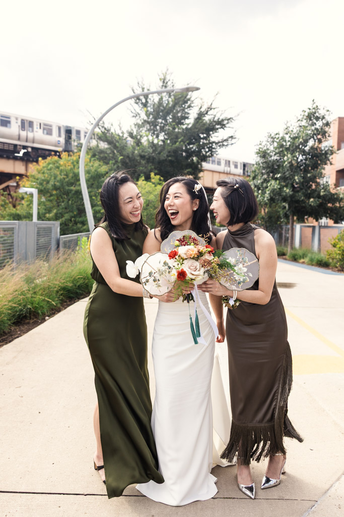 Bride and her bridesmaids in dresses laugh together outdoors, holding flower bouquets with a CTA train in the background before wedding celebration at The Joinery in Chicago