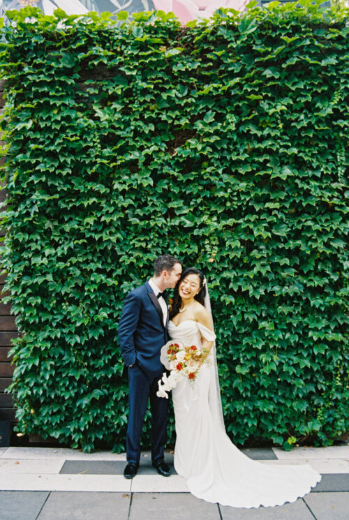 Bride and groom smiling and standing together in front of a lush green ivy-covered wall, their hands gently intertwined like seamless joinery.