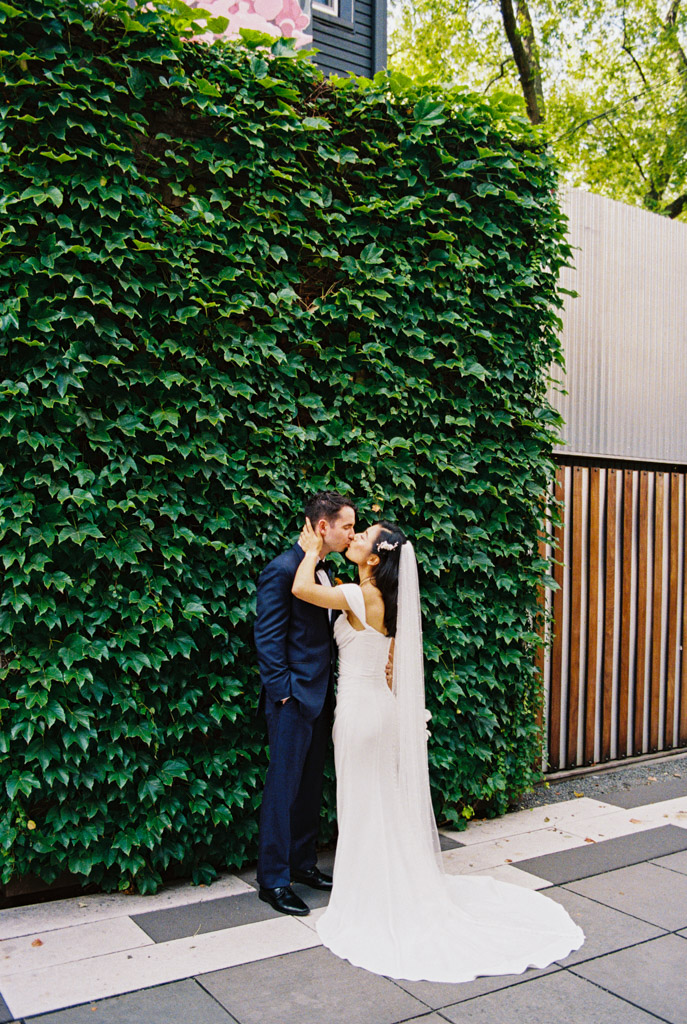 Bride and groom share a kiss in front of a leafy green wall and wooden fence outside the Joinery wedding venue in Chicago