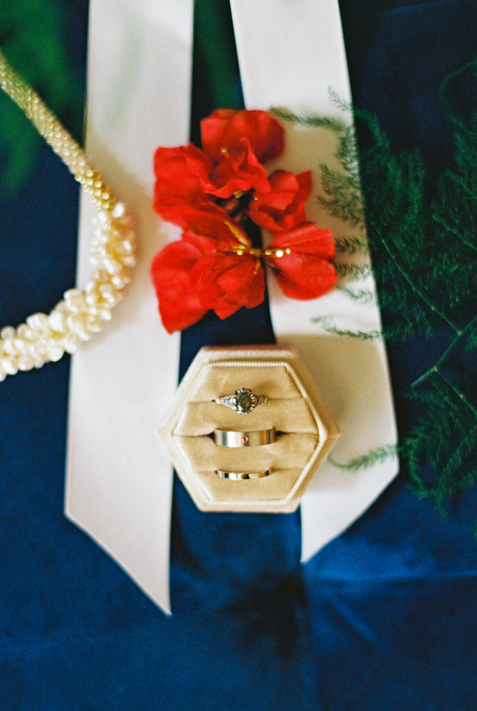 Wedding rings in a hexagonal box, red flowers, white ribbon, pearl necklace, and greenery arranged on dark blue fabric