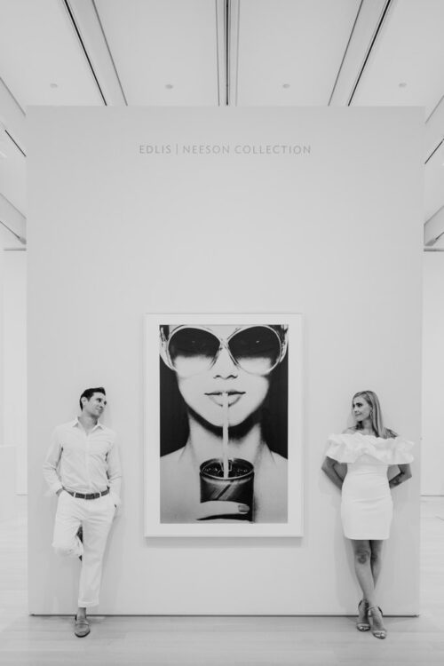 A man and woman in white outfits stand on either side of a large black-and-white portrait in an art gallery.