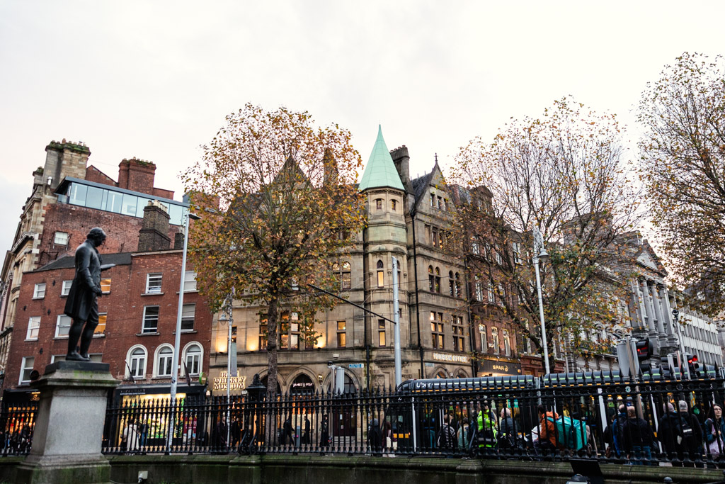 Crowds walk past historic buildings and trees in Dublin City Centre with a statue in the foreground