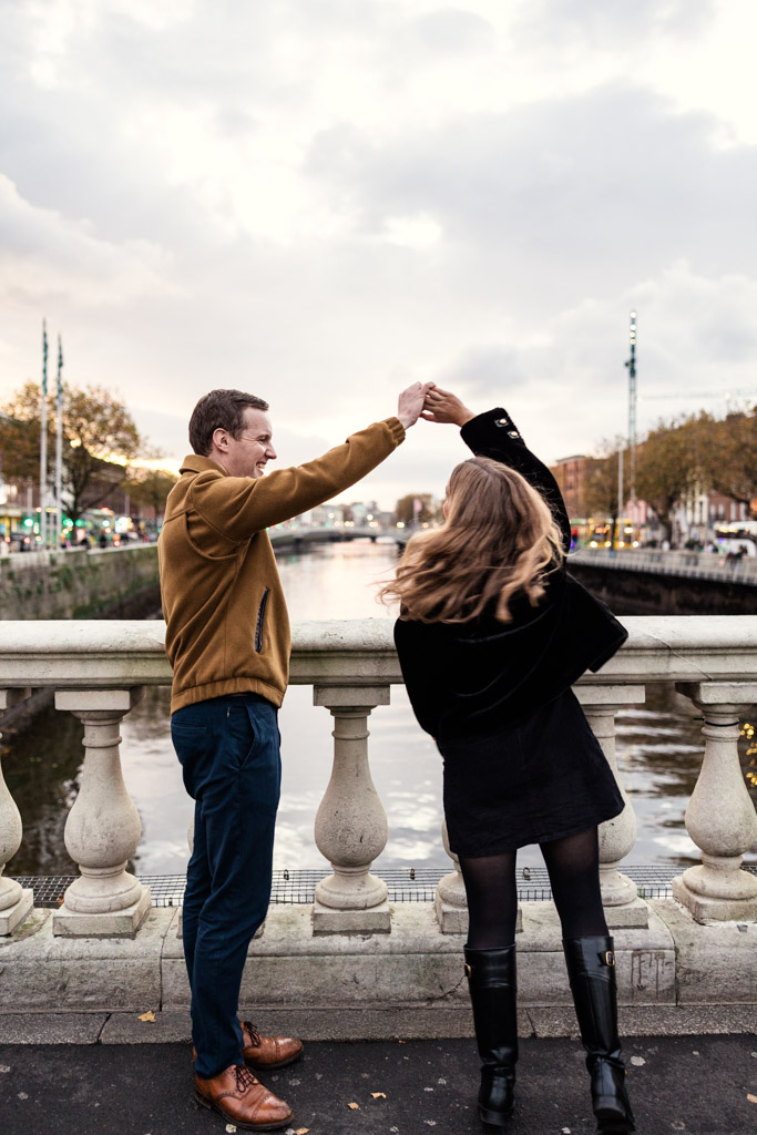 Man spins his partner as they stand on a Dublin bridge overlooking the River Liffey, with vibrant autumn trees in the background