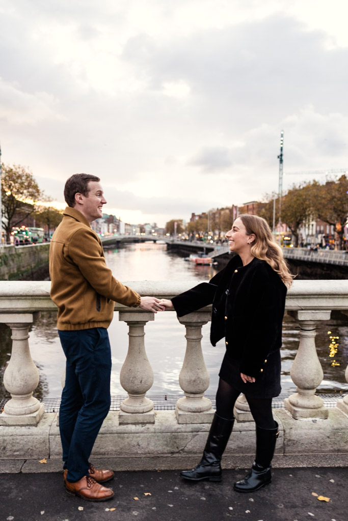 Man and woman hold hands and smile at each other on a bridge over the River Liffey at sunset during their Dublin engagement session