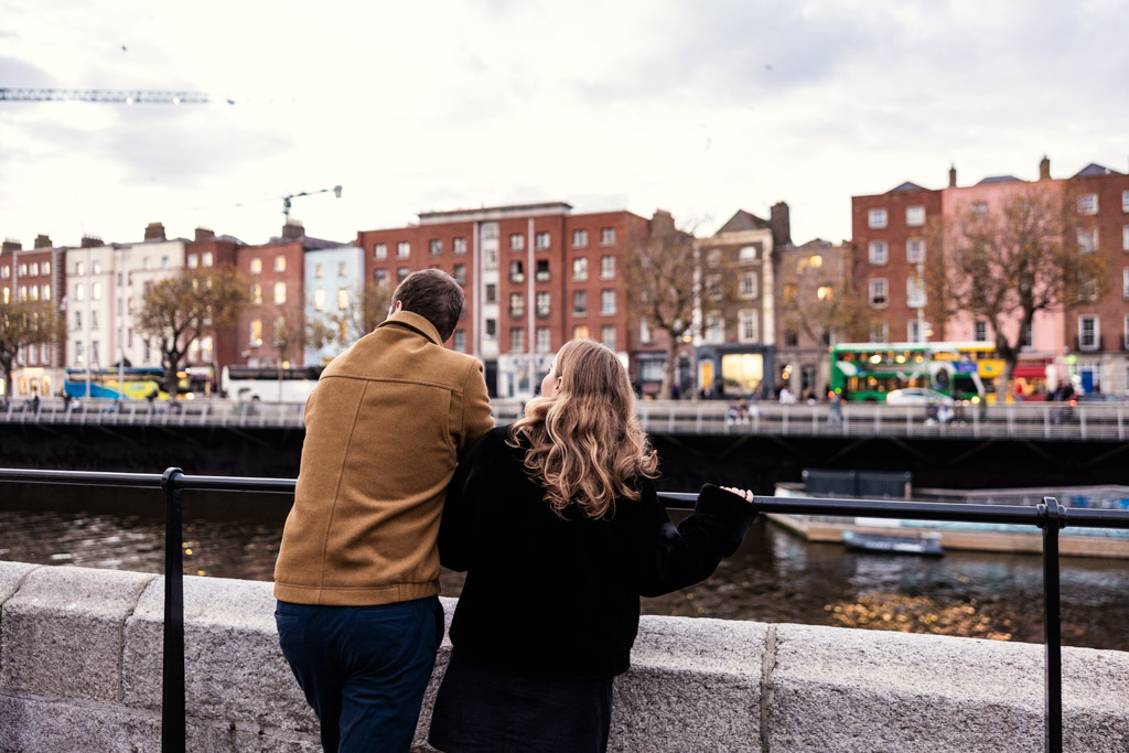 Engaged couple stands by a river, looking at Dublin city buildings on the opposite bank