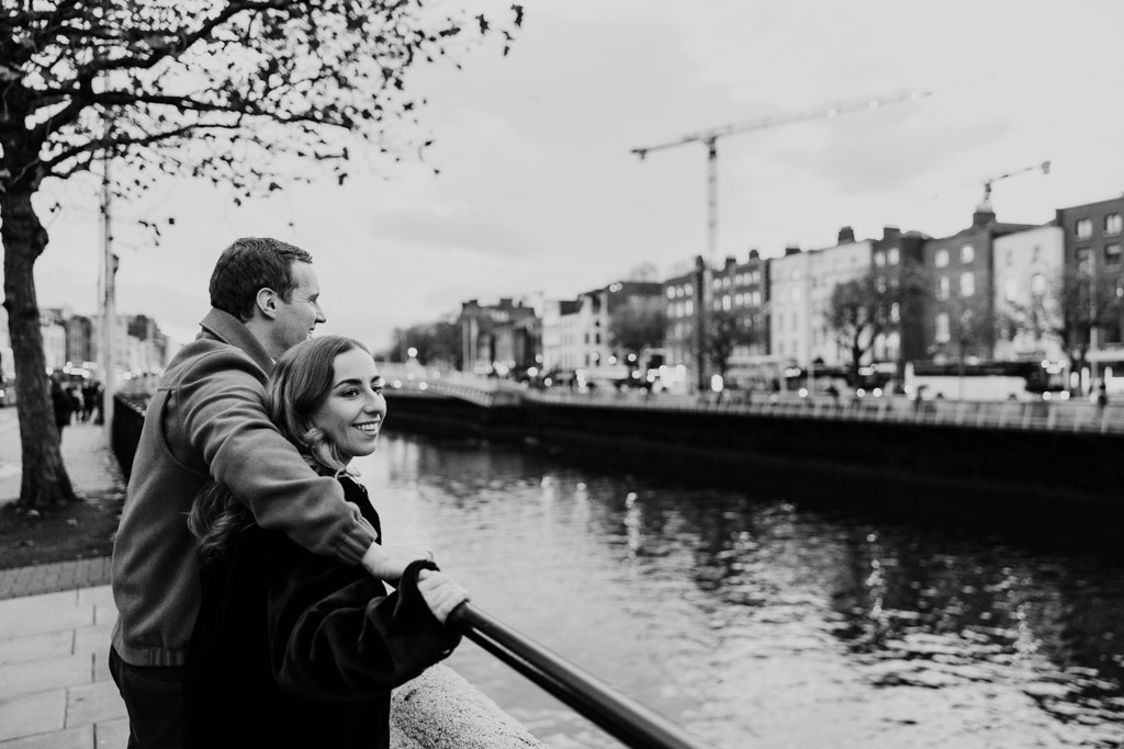Black and white photo of smiling couple standing by a river, enjoying an urban city view during their Dublin engagement session
