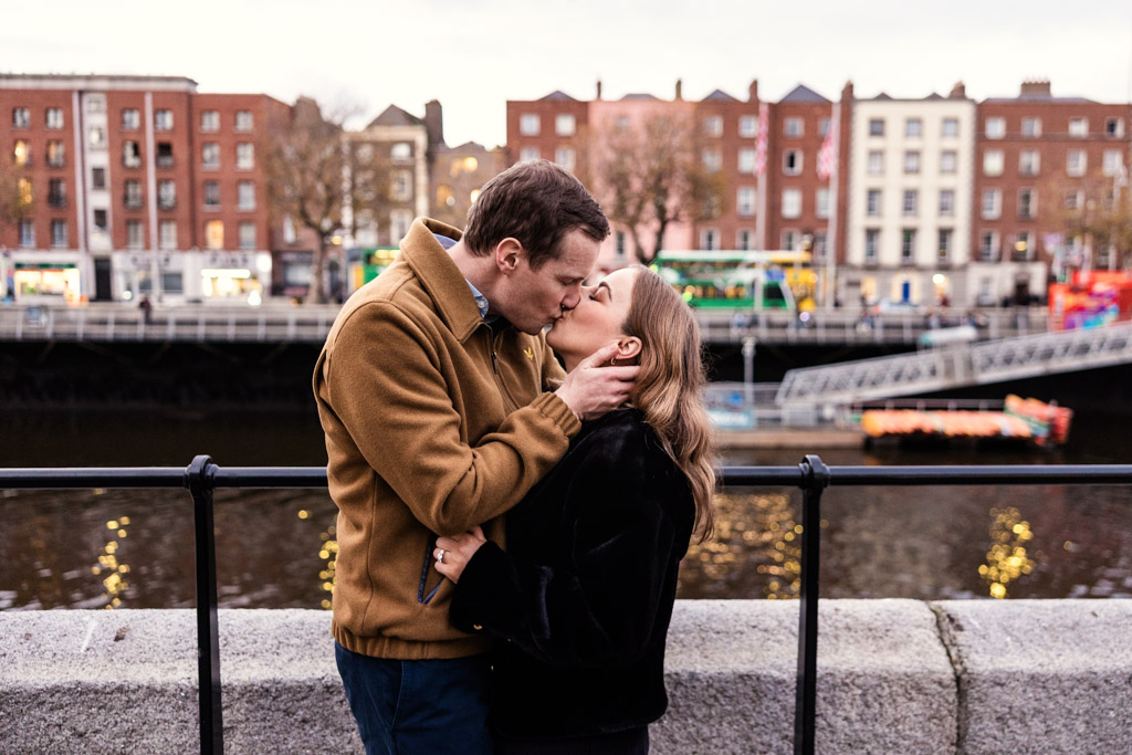 A couple shares a kiss by the riverside during their Dublin engagement, with city buildings and a passing tram in the background.