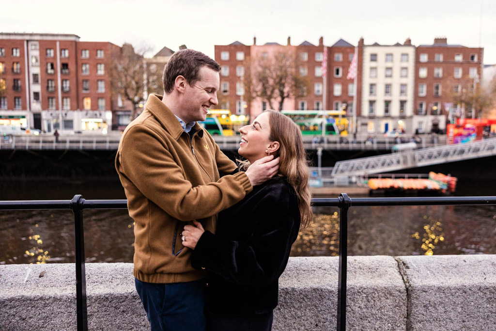 A couple smiles at each other while standing by a river, with city buildings in the background, capturing a romantic Dublin engagement moment
