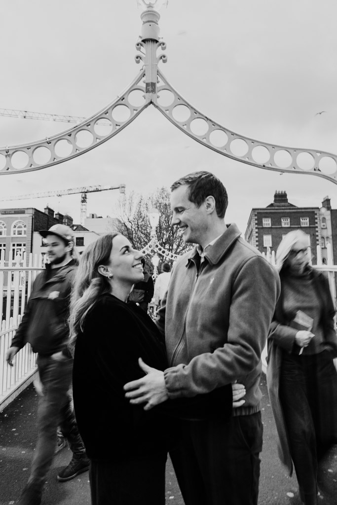 Black and white photo of happy couple under an ornate arch during their Dublin engagement session, as people walk by on the bustling city street