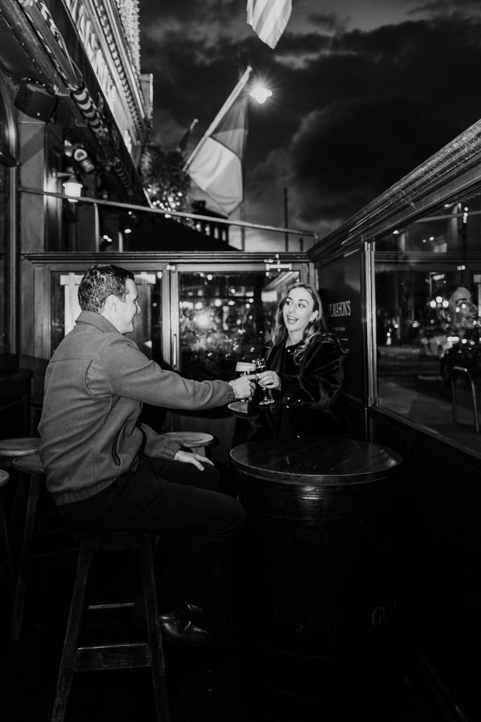Black and white photo of man and woman sitting outside at a bar in Dublin, clinking glasses and smiling at each other during their engagement session