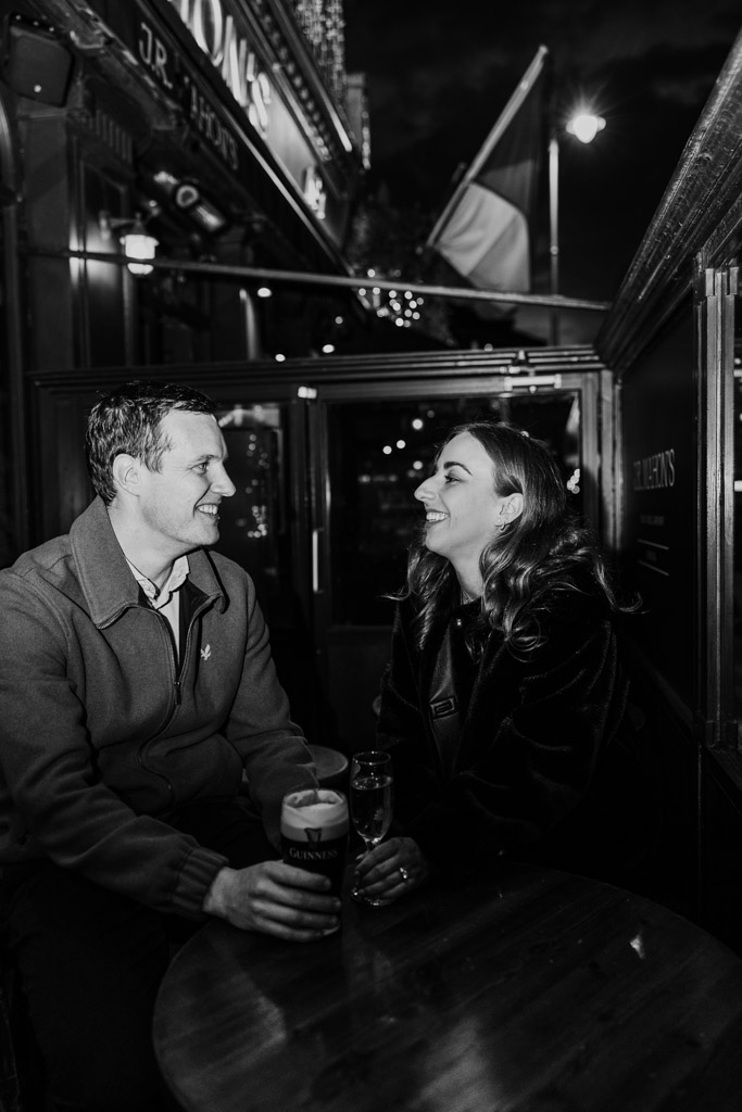 Black and white photo of man and woman smiling at each other while sitting outside a pub with drinks at night during their Dublin engagement session