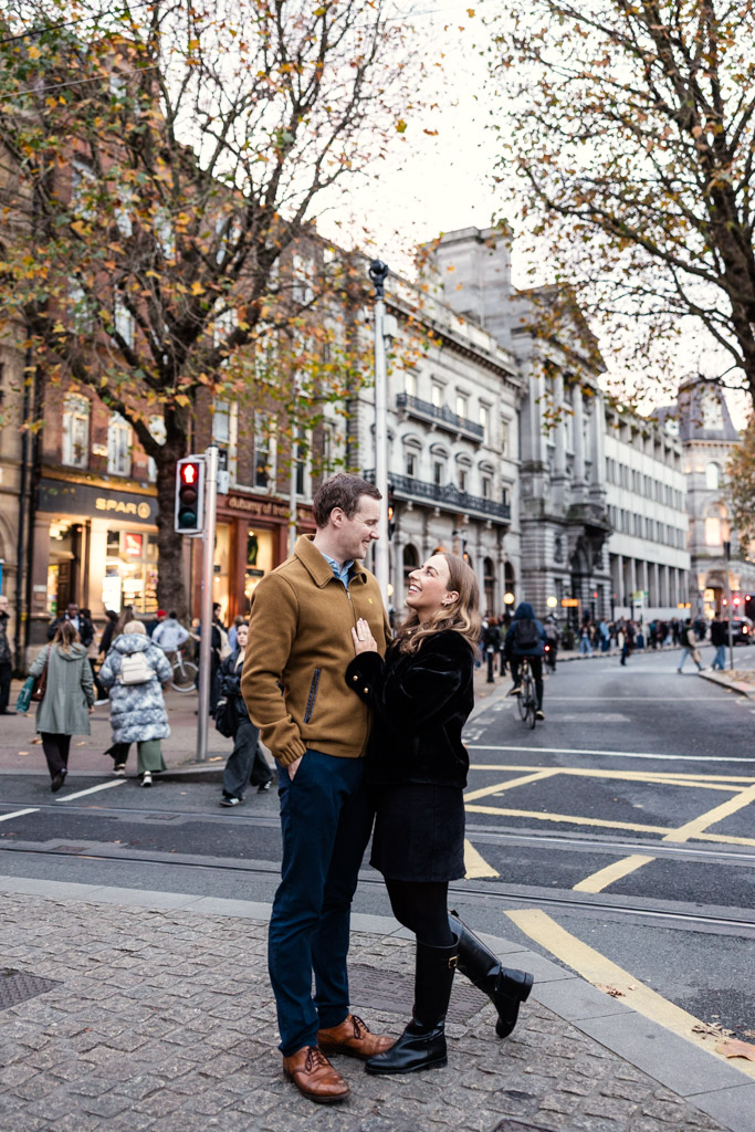 Engaged couple stands smiling at each other on a busy Dublin street corner, amid bustling people and iconic city buildings in the background