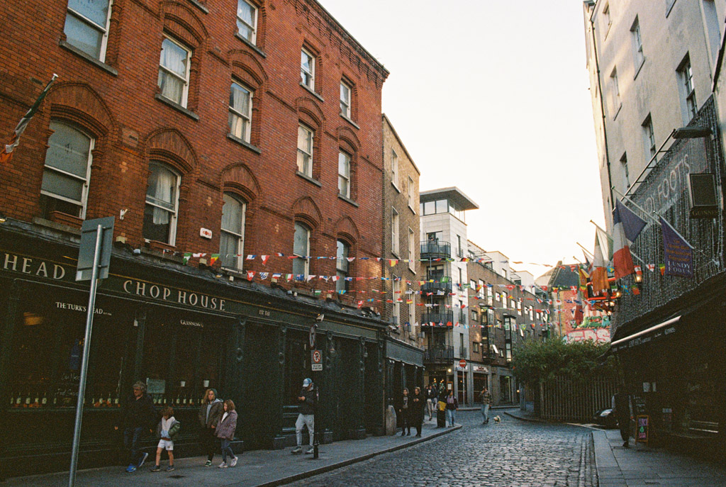 Film photo of a cobblestone street in Dublin lined with brick buildings and colorful bunting, bustling with people and shops