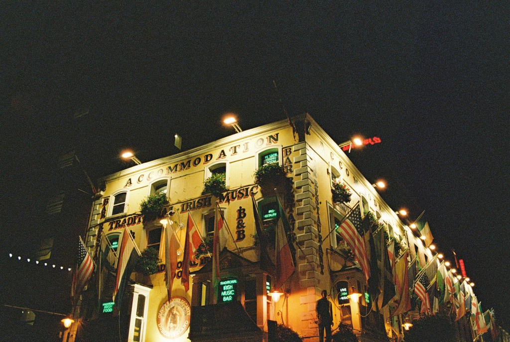 A brightly lit Dublin pub at night, decorated with flags and signs for accommodation and a music bar