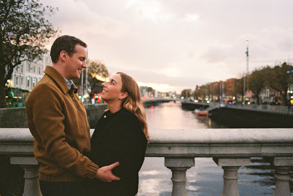 Film photo of engaged couple standing on a bridge at sunset during their Dublin engagement session, smiling at each other with a river and cityscape in the background