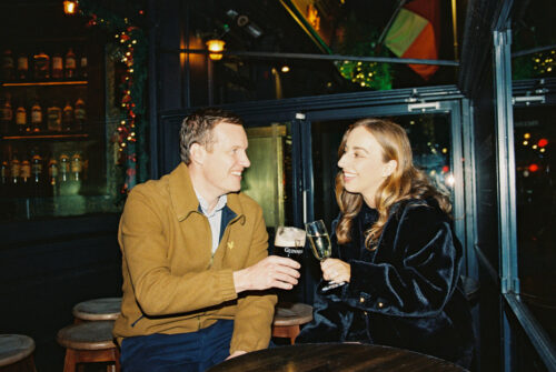 A man and woman smiling and clinking drinks at a cozy bar decorated with string lights, celebrating their Dublin engagement.
