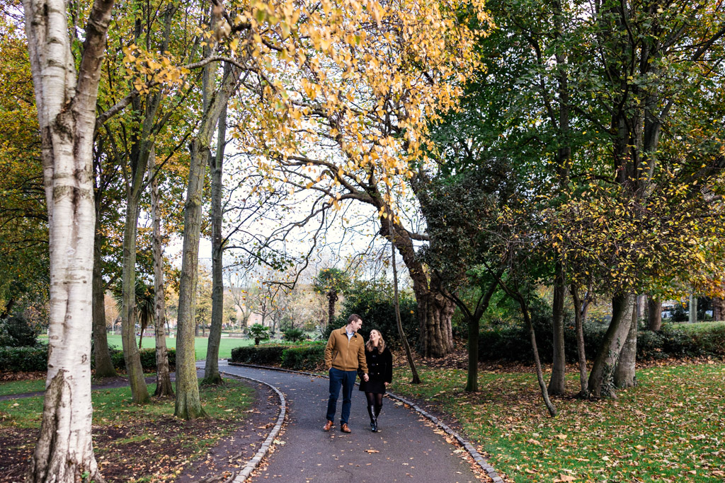 A couple enjoys a romantic stroll on a curved path in a park with autumn trees and fallen leaves, during their Dublin engagement session