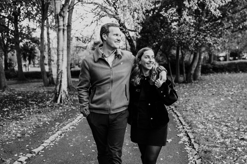 A couple walks arm in arm down a tree-lined path, smiling and enjoying a stroll in the park during their Dublin engagement.