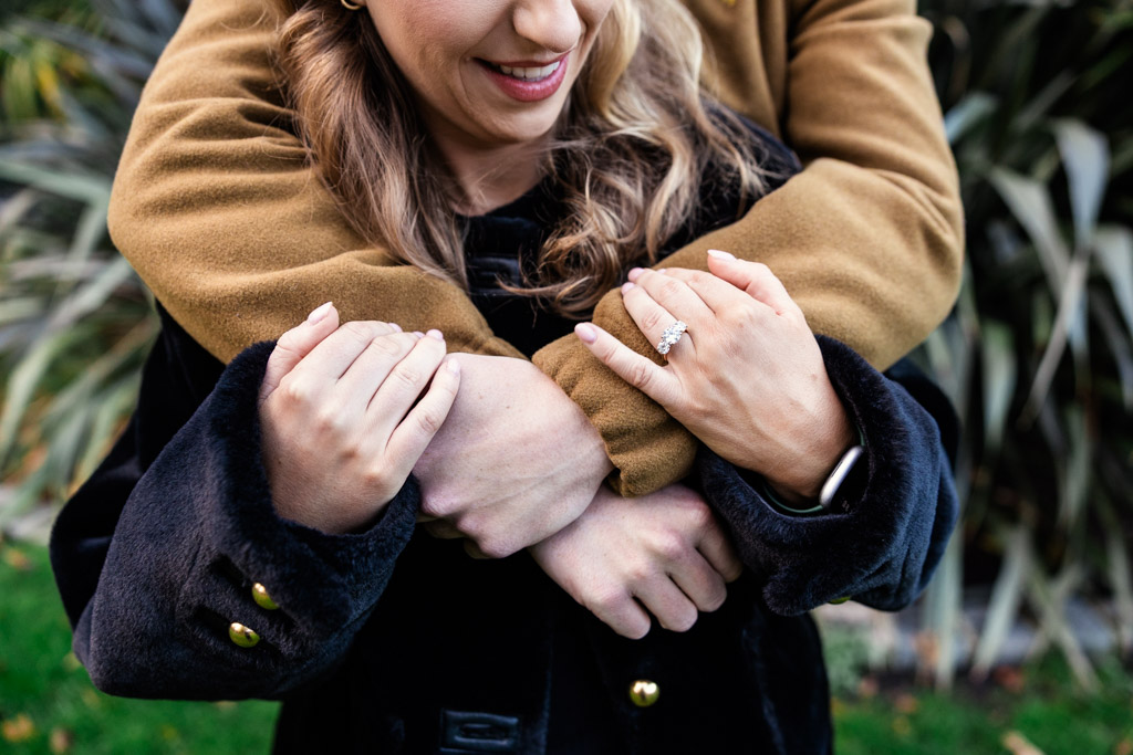 A woman smiles as her partner hugs her from behind, displaying her diamond engagement ring on her hand during Dublin engagement session