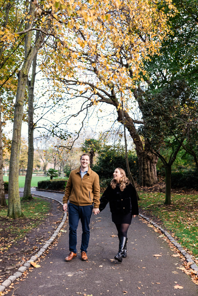 Engaged couple holding hands and smiling, walking on a park path lined with autumn trees in Merrion Square, Dublin