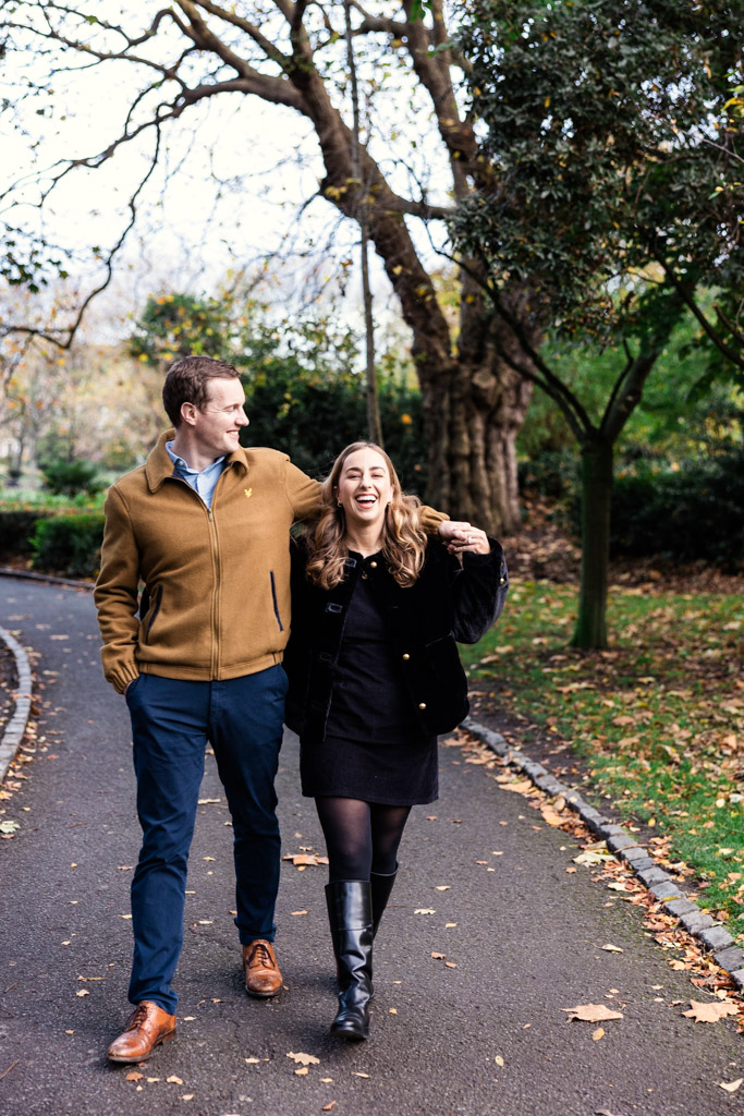 A man and woman walk arm in arm, smiling, on a tree-lined path in Merrion Square with autumn leaves during their Dublin engagement session