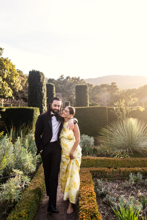 A couple walks in a sunny garden, the man in a tuxedo and the woman in a yellow floral dress, smiling.