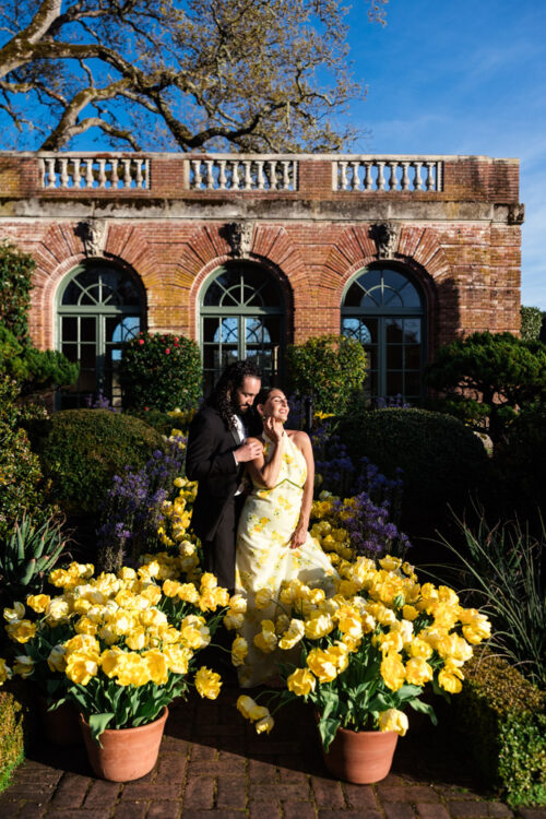 A couple in formal attire poses among yellow flowers in front of an elegant brick building.