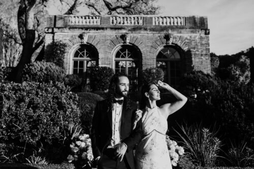 A couple in formal attire poses in front of a grand building and lush garden, in black and white.