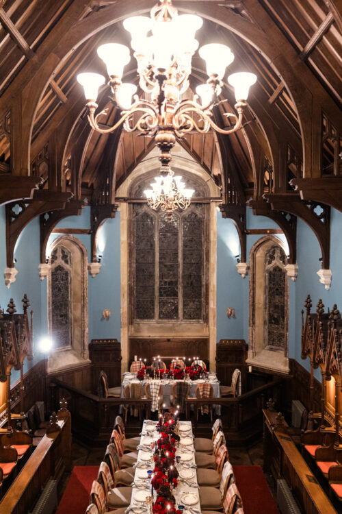 A long, elegant dining table is set in a grand, wood-paneled hall with chandeliers and tall arched windows—an idyllic setting for an unforgettable Ireland wedding.