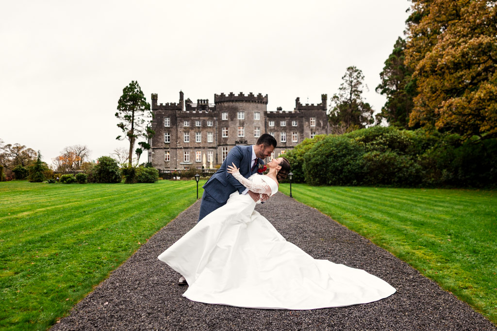 A groom dips his bride in a white dress on a path in front of Markree Castle