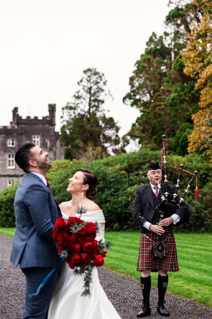 Bride and groom smile while a man in a kilt plays bagpipes with Markree Castle and trees in the background